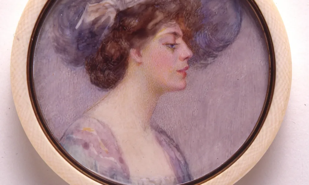 A miniature portrait in a round, white frame of a light-skinned adult woman in profile with brown hair pulled back in a loose bun. She wears a large, periwinkle hat that matches the sleeves of her purple and blue dress. The background of the portrait is pale purple.