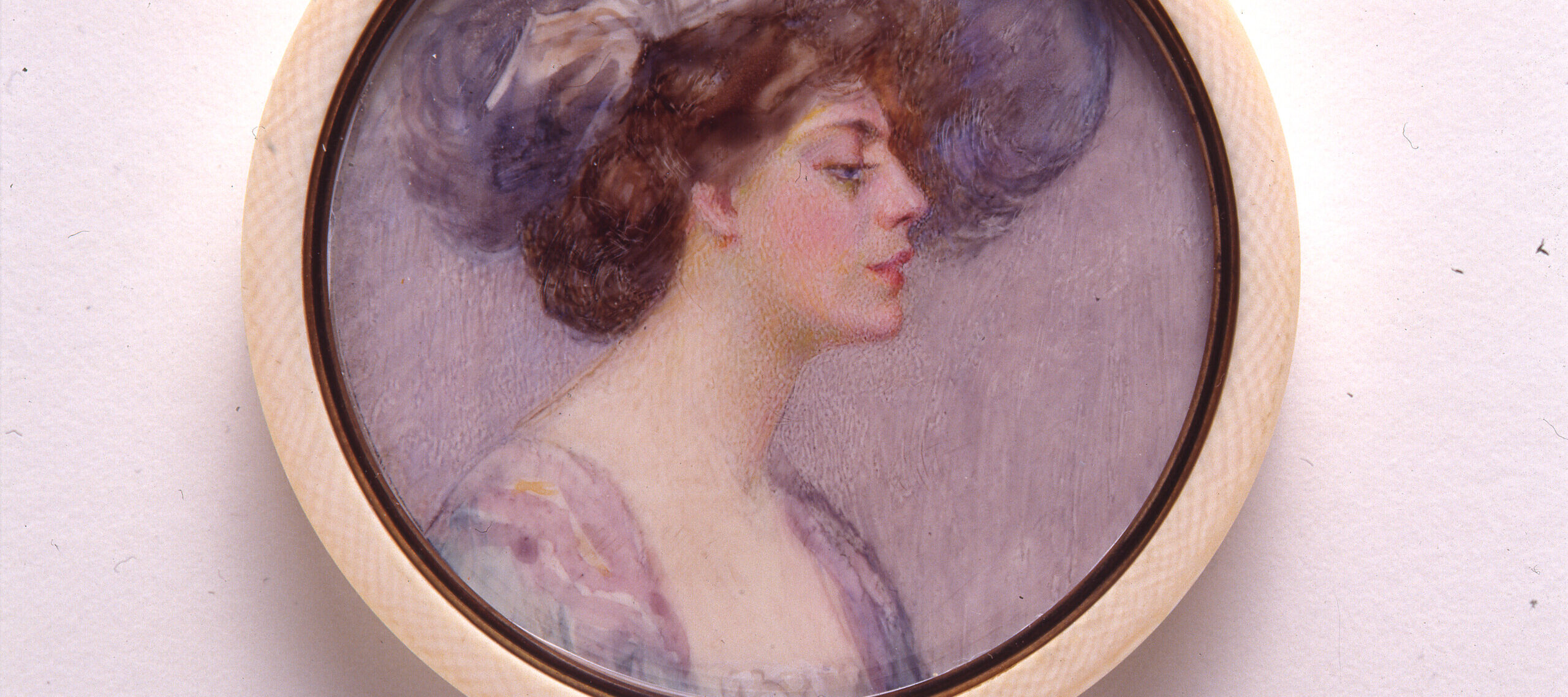 A miniature portrait in a round, white frame of a light-skinned adult woman in profile with brown hair pulled back in a loose bun. She wears a large, periwinkle hat that matches the sleeves of her purple and blue dress. The background of the portrait is pale purple.