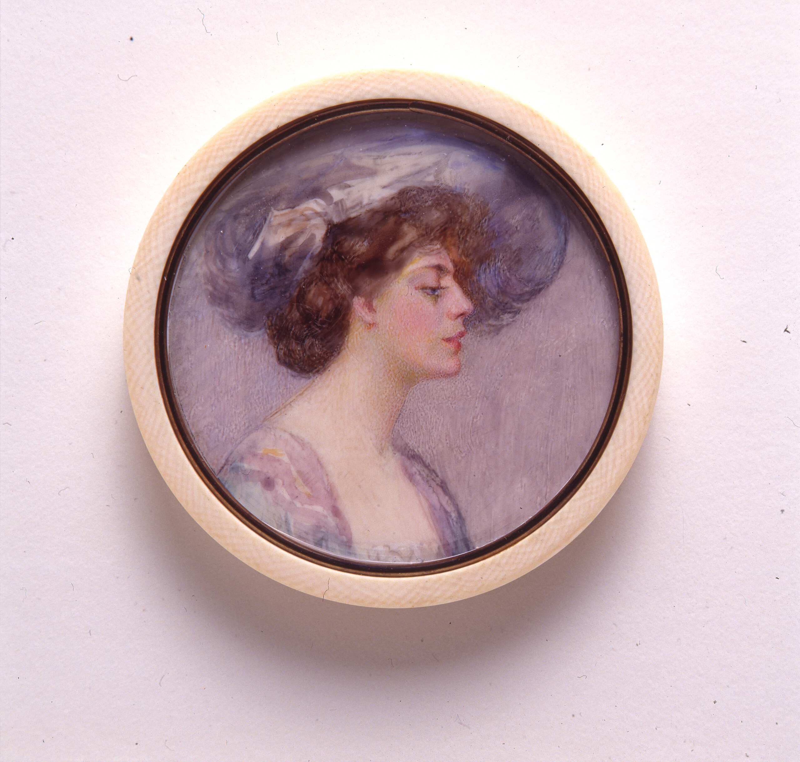 A miniature portrait in a round, white frame of a light-skinned adult woman in profile with brown hair pulled back in a loose bun. She wears a large, periwinkle hat that matches the sleeves of her purple and blue dress. The background of the portrait is pale purple.