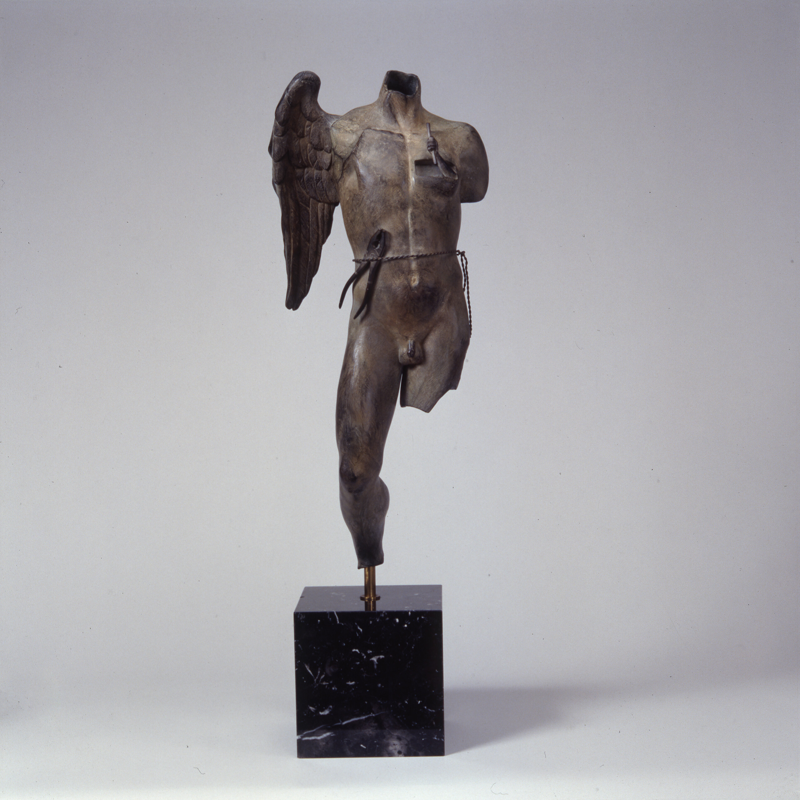 A bronze statue of a man's torso with one leg and one wing. The other leg is broken off. There is a thread around his waist, holding tongs. The broken-off leg and body make the sculpture seem ancient, as if it was retrieved from the ruins of an ancient city.