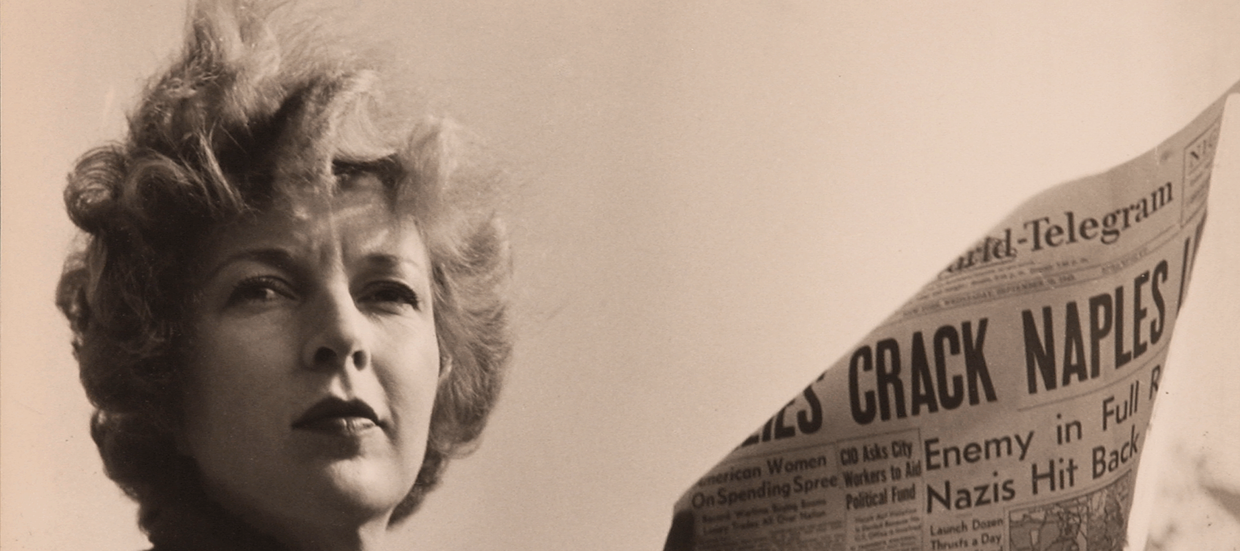A black-and-white photograph of a light-skinned adult woman holding a newspaper with news about World War II. She wears a coat and her short, curly hair is caught in the wind.