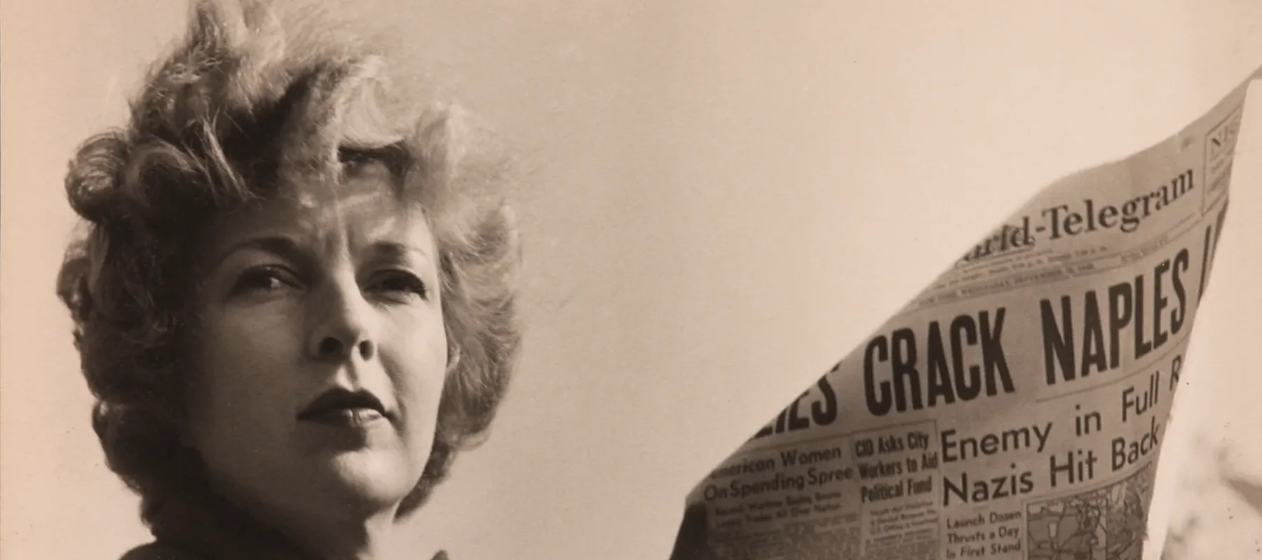 A black-and-white photograph of a light-skinned adult woman holding a newspaper with news about World War II. She wears a coat and her short, curly hair is caught in the wind.