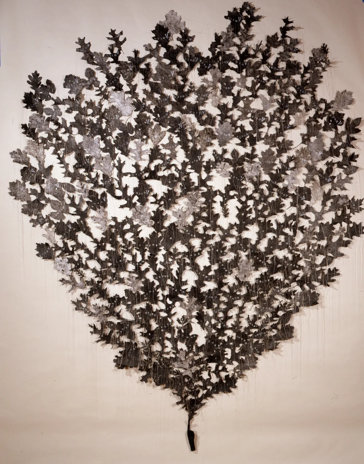 Hundreds of light- and dark-gray shapes resembling oak leaves are mounted directly to a wall in the shape of an ice-cream cone. They appear to sprout from the index finger of a small, coal-black hand mounted near the floor. Delicate strands of thread hang down from the leaves.