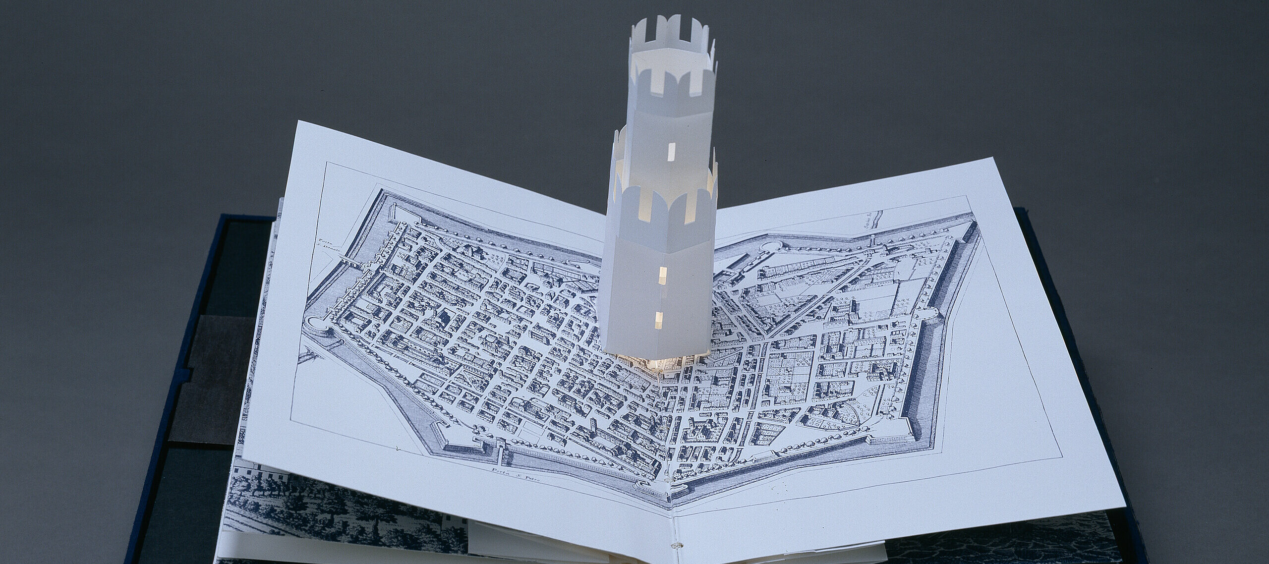 A pop-up book displays an intricate paper model of a tower rising from a map illustration of an old city, all set against a dark gray background.