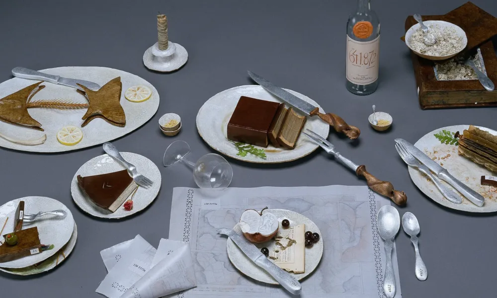 A photo of a still life of the remnants of a large meal. The half-eaten food is actually made of books that are sculpted to resemble fishbones, slices of pie, and other food scraps.