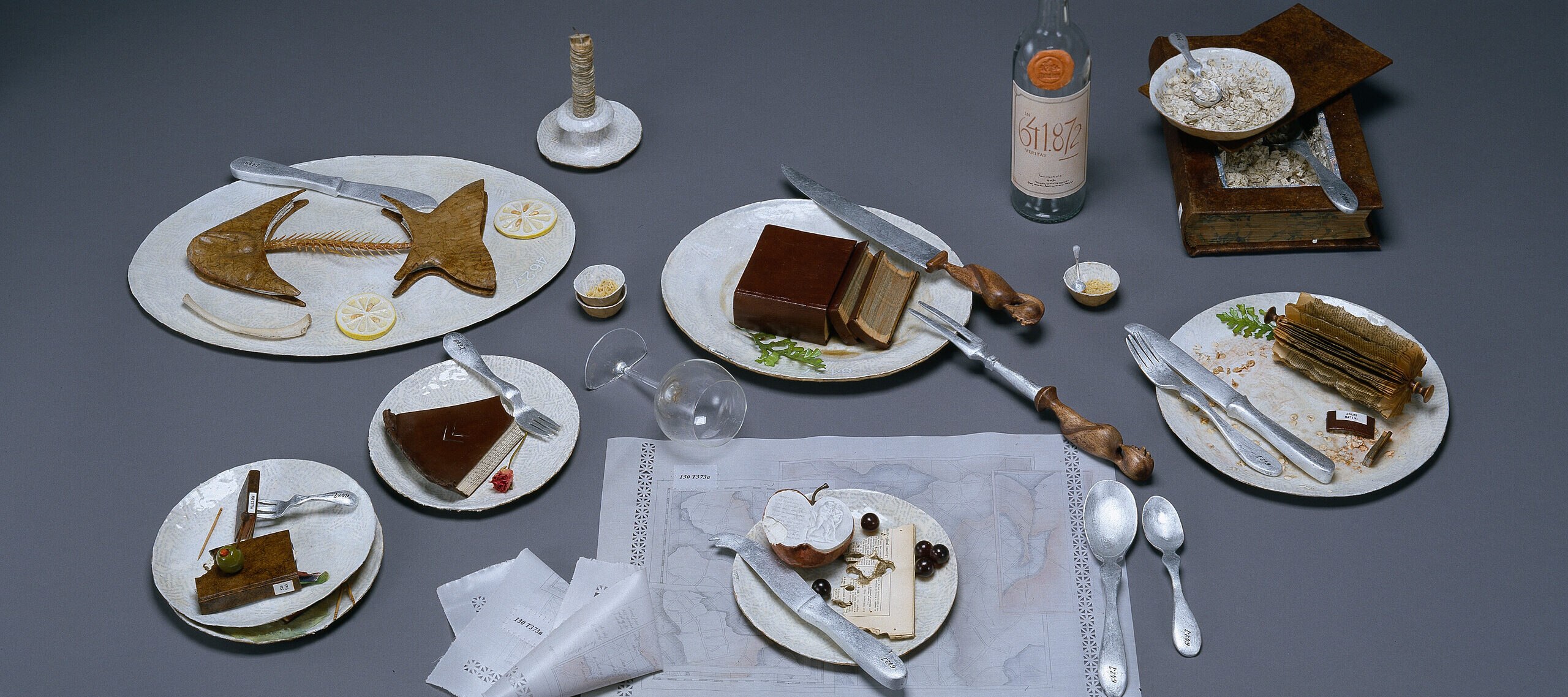 A photo of a still life of the remnants of a large meal. The half-eaten food is actually made of books that are sculpted to resemble fishbones, slices of pie, and other food scraps.
