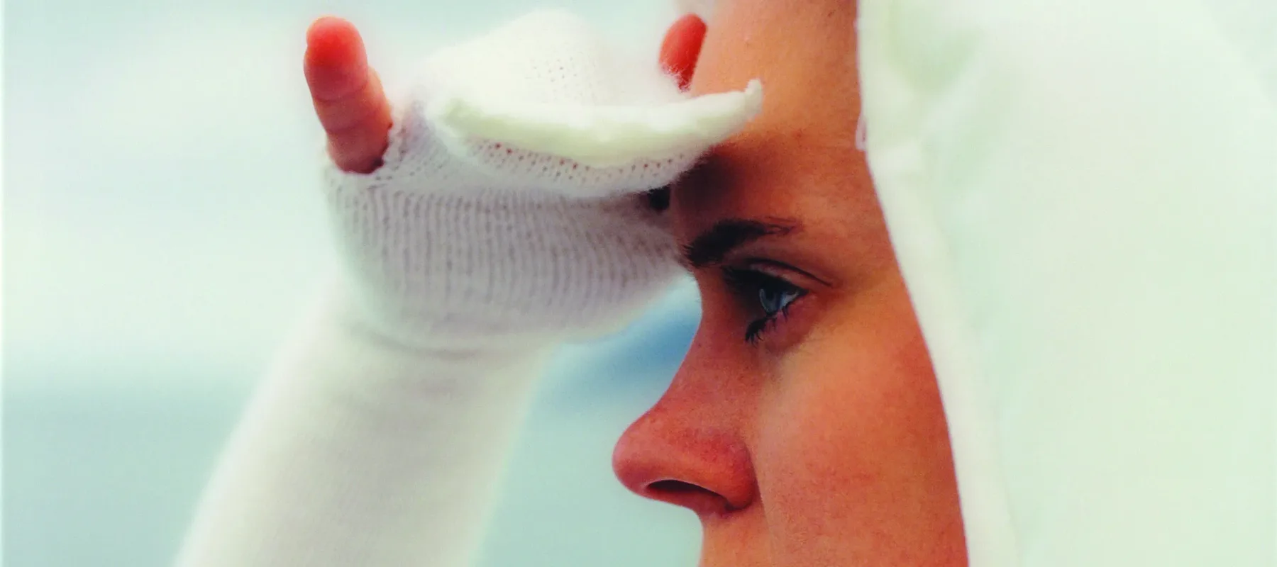 Square close-up of a light-skinned woman in profile looking to the left, seen from the shoulders up. She wears all white, including a bonnet and knit sleeves that become gloves covering her three middle fingers, and she holds her right hand up as if shielding her eyes from the sun.