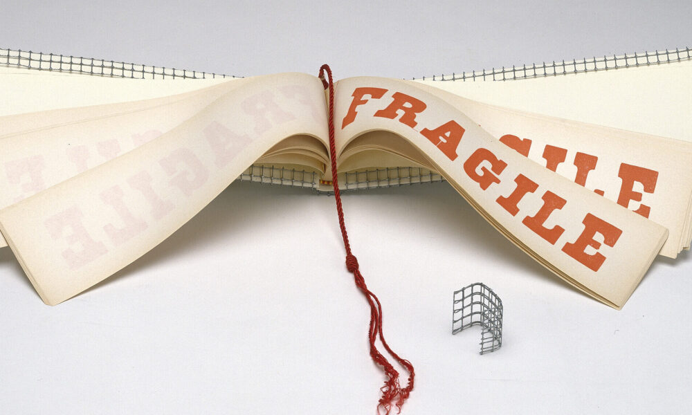 A wide and narrow book opened to display pages with the word FRAGILE in red block letters. A red tassel hangs down the center of the book and the cover is wrapped with wire mesh.