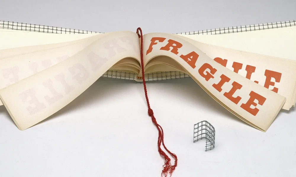 A wide and narrow book opened to display pages with the word FRAGILE in red block letters. A red tassel hangs down the center of the book and the cover is wrapped with wire mesh.