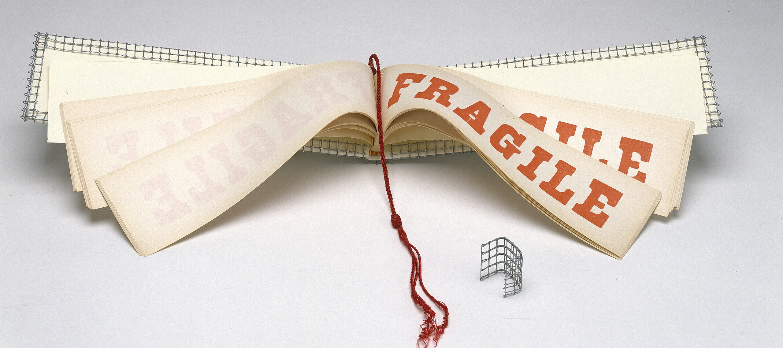 A wide and narrow book opened to display pages with the word FRAGILE in red block letters. A red tassel hangs down the center of the book and the cover is wrapped with wire mesh.