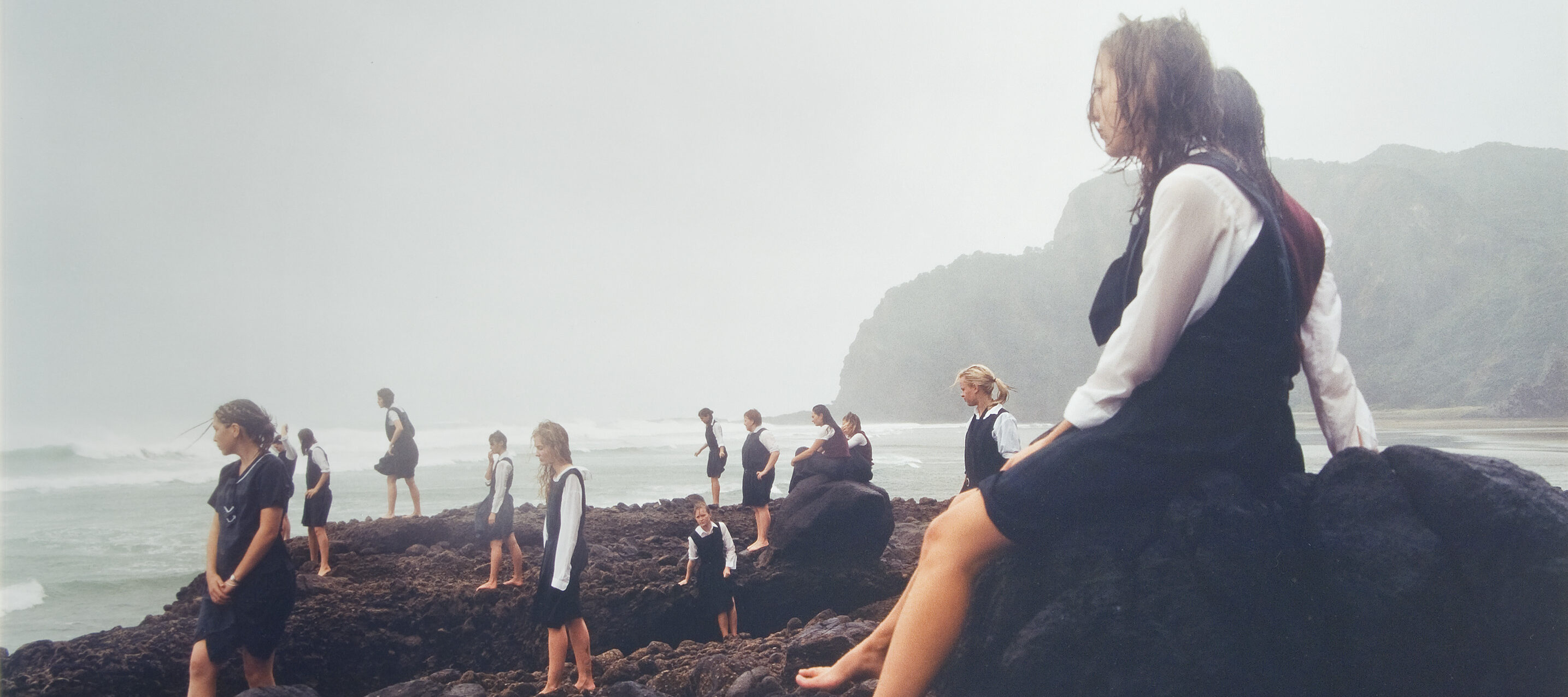 Fifteen light-skinned teenage girls stand and sit on dark brown, rocky outcropping. They face left, in profile, out toward stormy waves, and they wear the same school uniform of long-sleeved, white, collared shirts and navy blue dresses. They are soaked by the storm and barefoot.