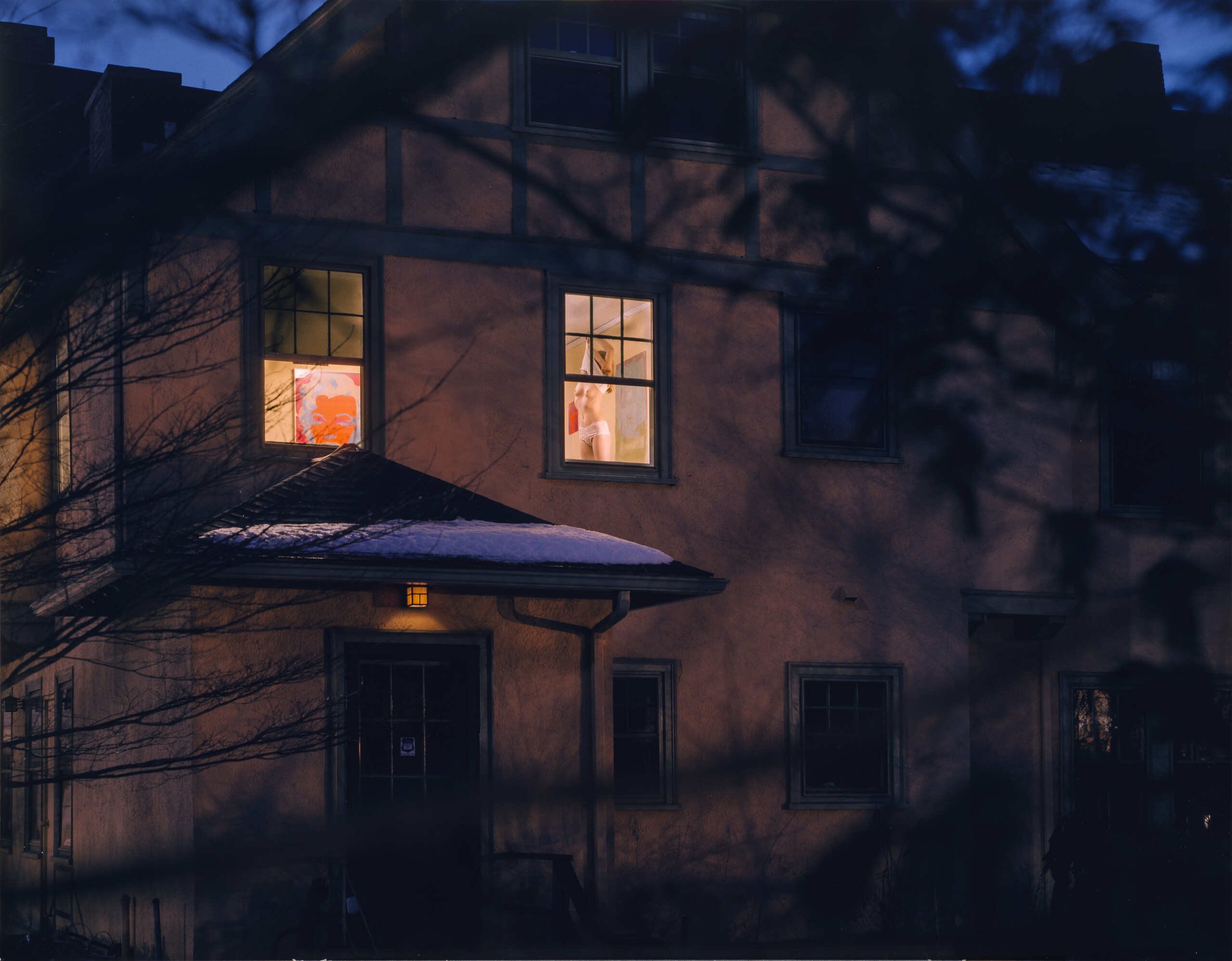 The view of a two-story house at night. The top room of the house has its lights on and you can see through the window the silhouette of a girl pulling a shirt over her head and exposing her torso. In the forefront of the photo, tree branches create shadows and frame the window.