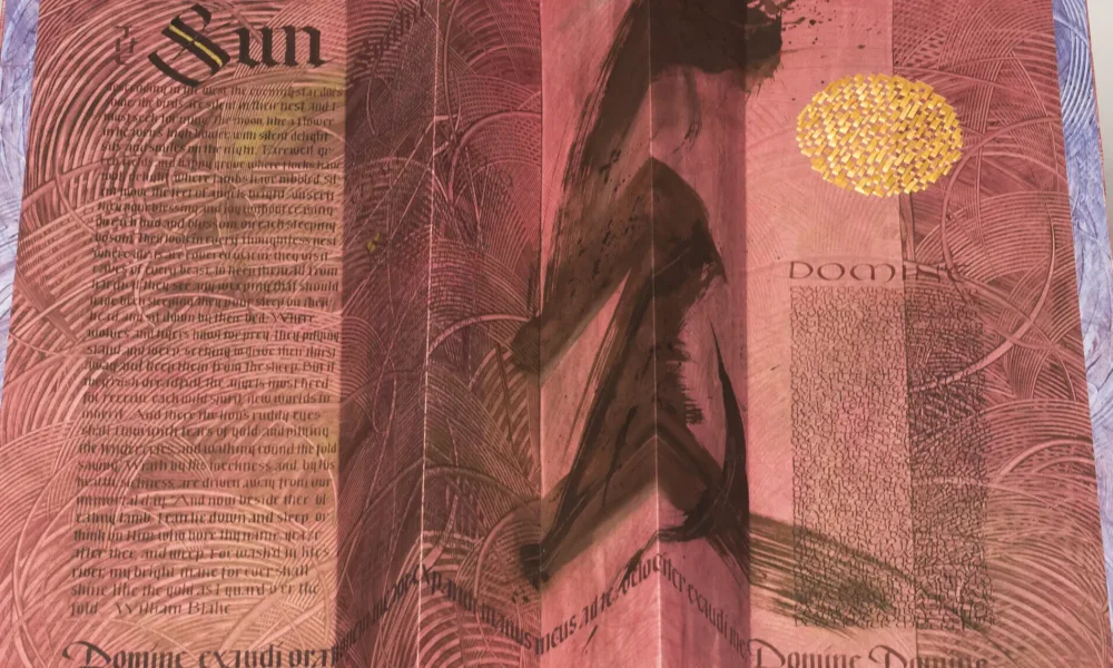 An open, accordion-style book with red-pink pages and black text. The word "Sun" is written in large, Gothic print at the upper left. At center-right is a large, black brushstroke that extends the length of the page. At the upper right is a golden circle.