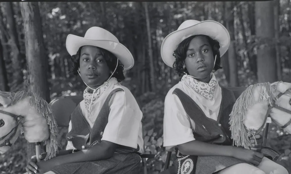 A black-and-white photograph of two dark-skinned young twins sitting in folding chairs against a woodsy background, facing the camera with their bodies angled outward in opposite directions. They wear matching cowgirl outfits, complete with hats, boots, and hobby horses.