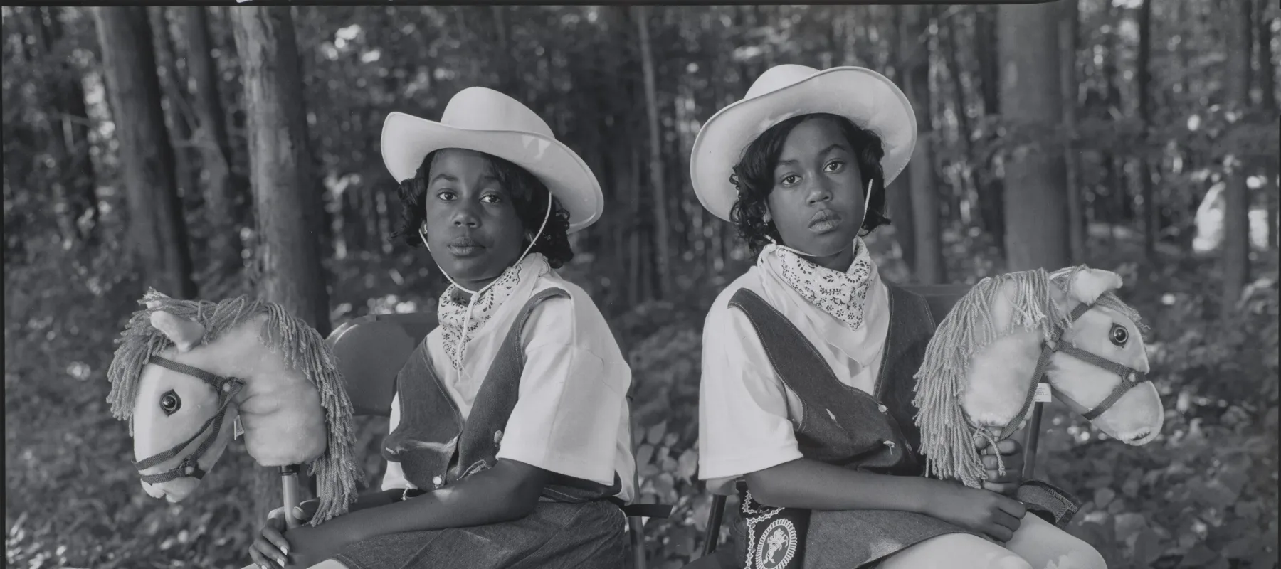 A black-and-white photograph of two dark-skinned young twins sitting in folding chairs against a woodsy background, facing the camera with their bodies angled outward in opposite directions. They wear matching cowgirl outfits, complete with hats, boots, and hobby horses.