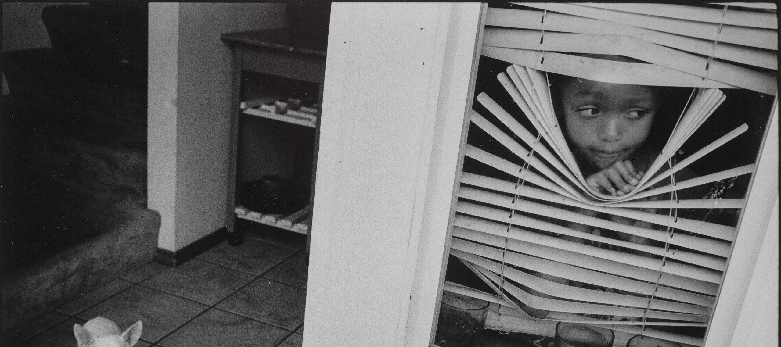 A black-and-white photograph of a young, medium-dark skinned girl peeking out between white blinds in a window that she bends and holds open with one hand. To the left of the window is an open doorframe to a tiled room where a little white dog stands.