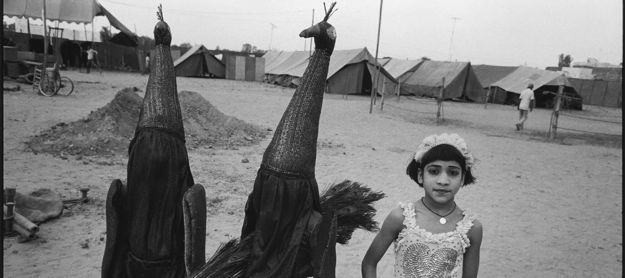 A black-and-white photograph of a medium-skinned girl in a frilly acrobat costume standing next to two children in identical, full-body peacock costumes. One peacock-costumed child holds a bundle of long peacock feathers, and they all stand on sand with a row of tents behind them.