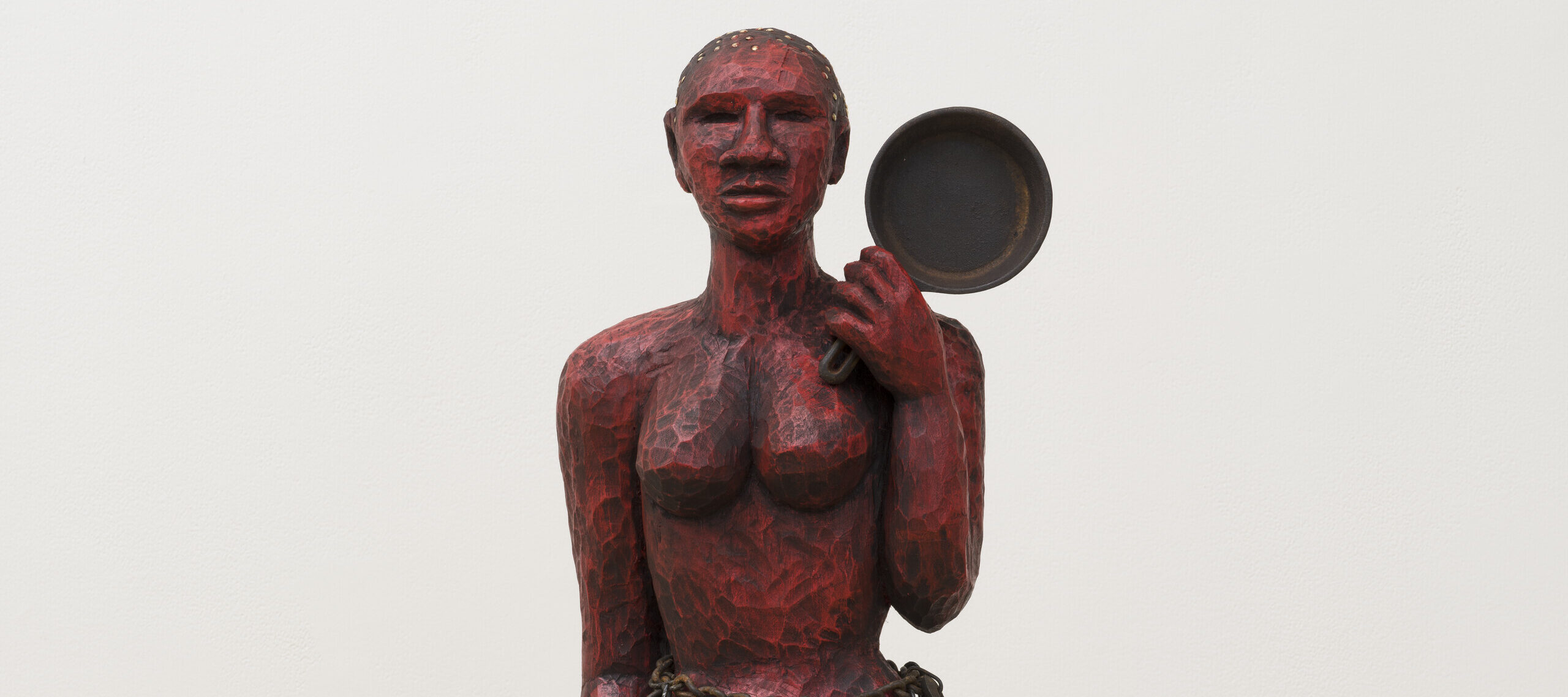 A red painted wooden sculpture of a woman holding a cast iron skillet in her left hand. She has a heavy chain around her waist with multiple skillets hanging from it like a skirt. She stands on a small wooden platform.