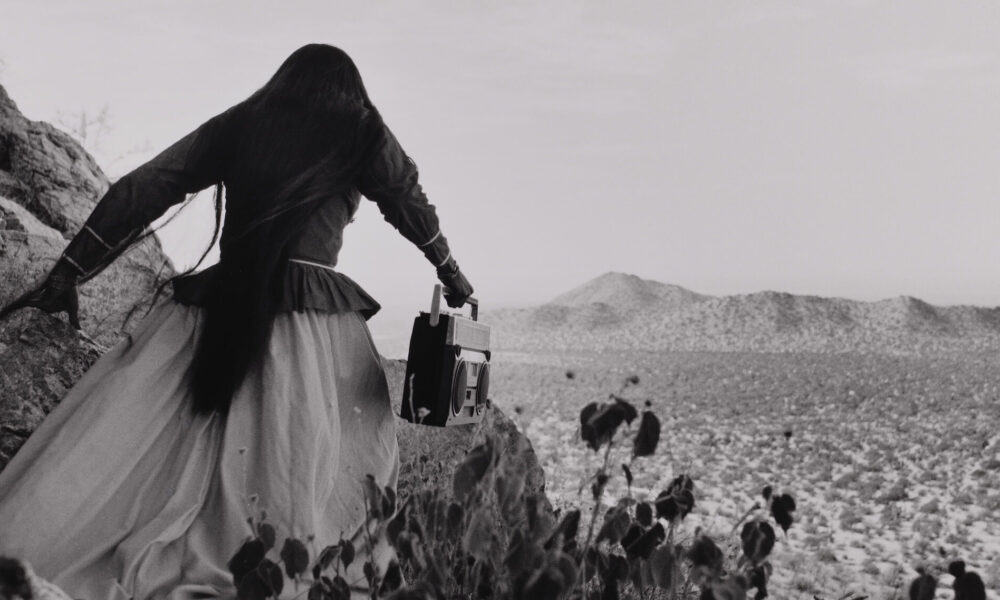 A black-and-white photograph shows the back of a woman as she crests a rocky path above a vast desert landscape beneath an expansive sky. Her traditional, ethnic full skirt, long-sleeved blouse, and long, straight, dark hair contrasts with the modern portable stereo she carries.