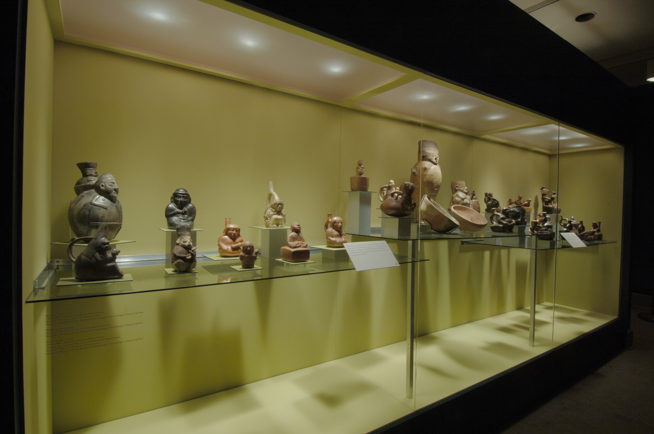 A view of a glass cabinet with little ancient clay sculptures inside. The cabinet is painted inside in a green color, which contrasts with the dark brown clay sculptures. The sculptures are of people, and there are also two bowls presented in the cabinet.