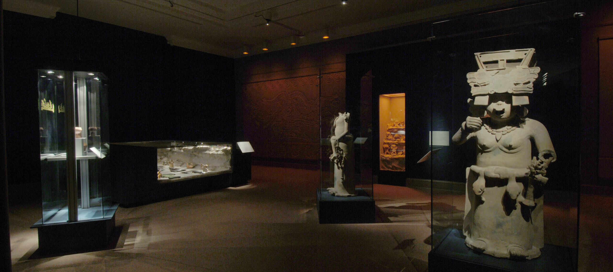 In a dark room, there are several artifacts standing on pedestals and sitting in glass cabinets. On the right, there is an ancient stone sculpture with a large headdress and snakes around their hip, presumably a god or goddess.