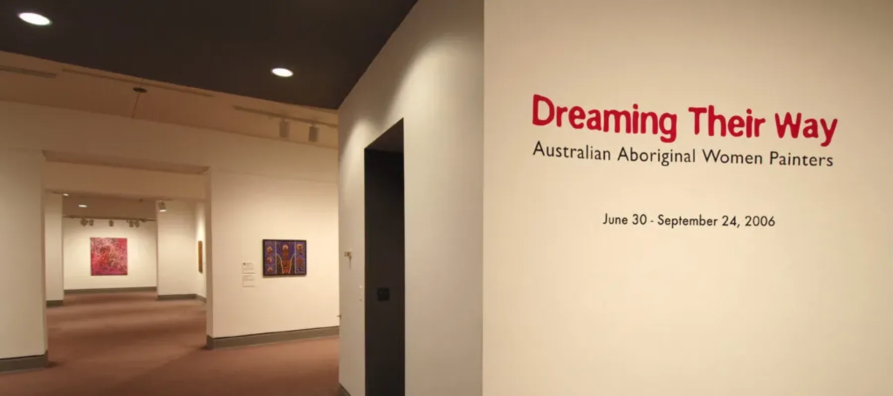 An installation view of a gallery space. There are paintings on the wall in the back, and on the wall in the front it says: "Dreaming Their Way" in big red letters, and "Australian Aboriginal Women Painters" in smaller, black letters.