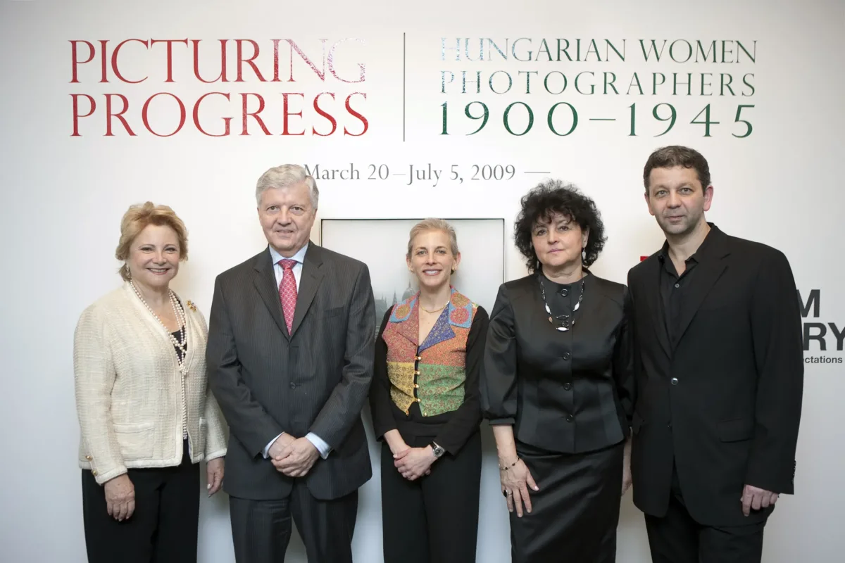 A group pf people with a light skin tone stand before a white wall. On the wall, in big red and green letters, it says: "Picturing Progress: Hungarian Women Photographers 1900–1945"