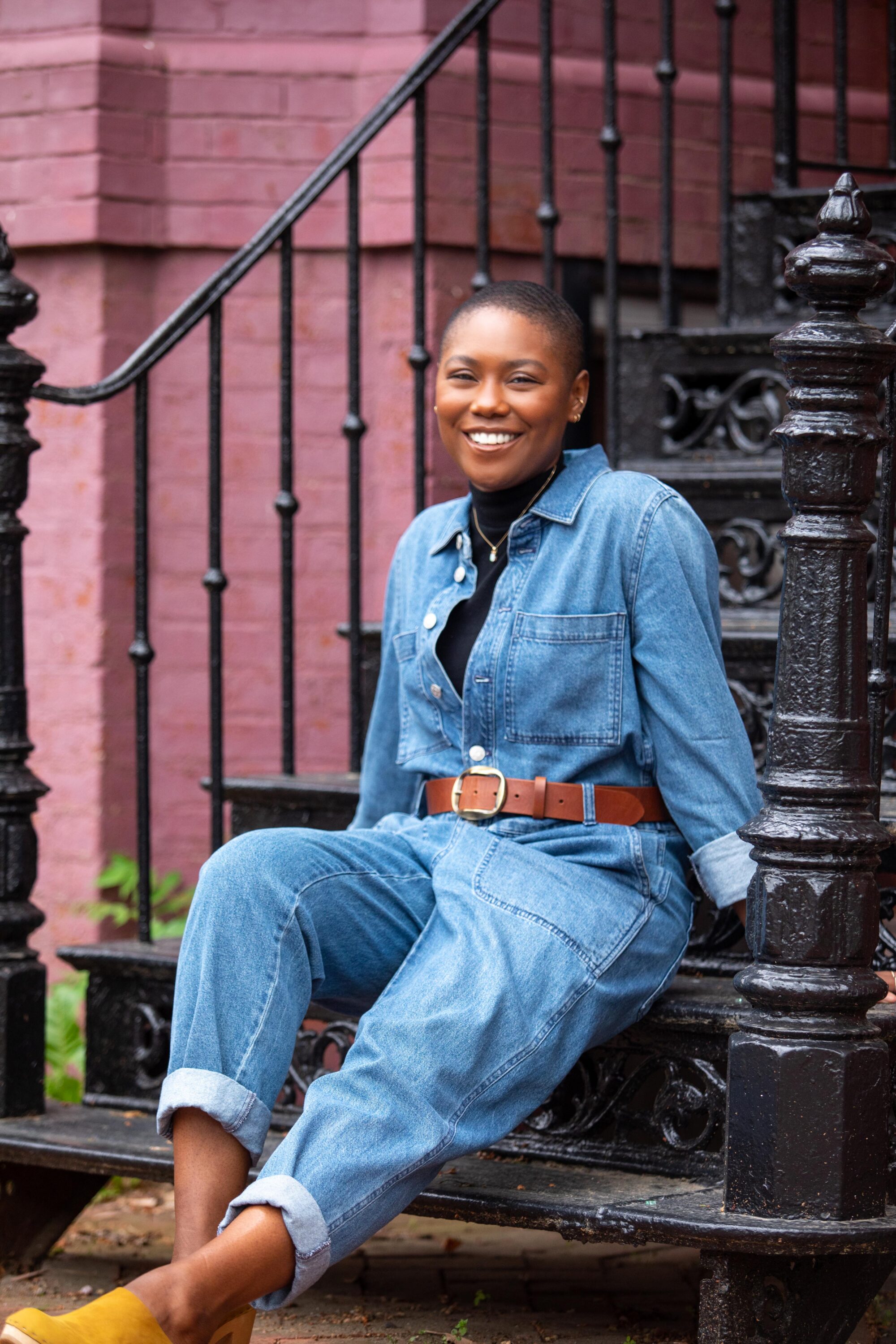 A youthful, dark-skinned woman sits casually on black iron stairs outside. She wears a denim jumpsuit, belted, with yellow clogs and smiles warmly at the camera. Her hair is worn short and close-cropped.
