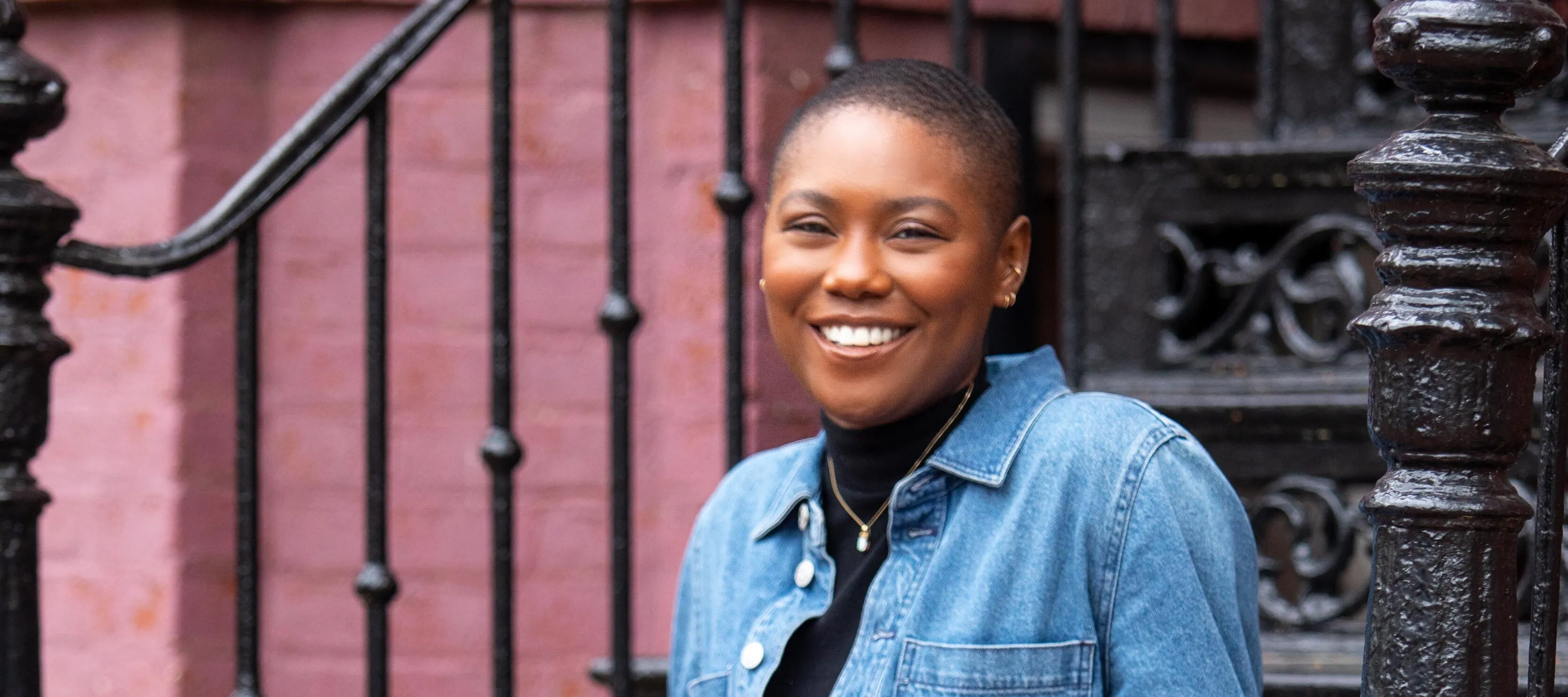 A youthful, dark-skinned woman sits casually on black iron stairs outside. She wears a denim jumpsuit, belted, with yellow clogs and smiles warmly at the camera. Her hair is worn short and close-cropped.