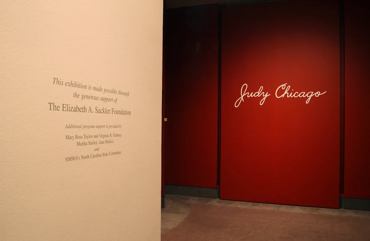 View of a gallery space. One wall is red, and in big letters, it says "Judy Chicago." On the opposite wall, the supporters of the exhibition are named.