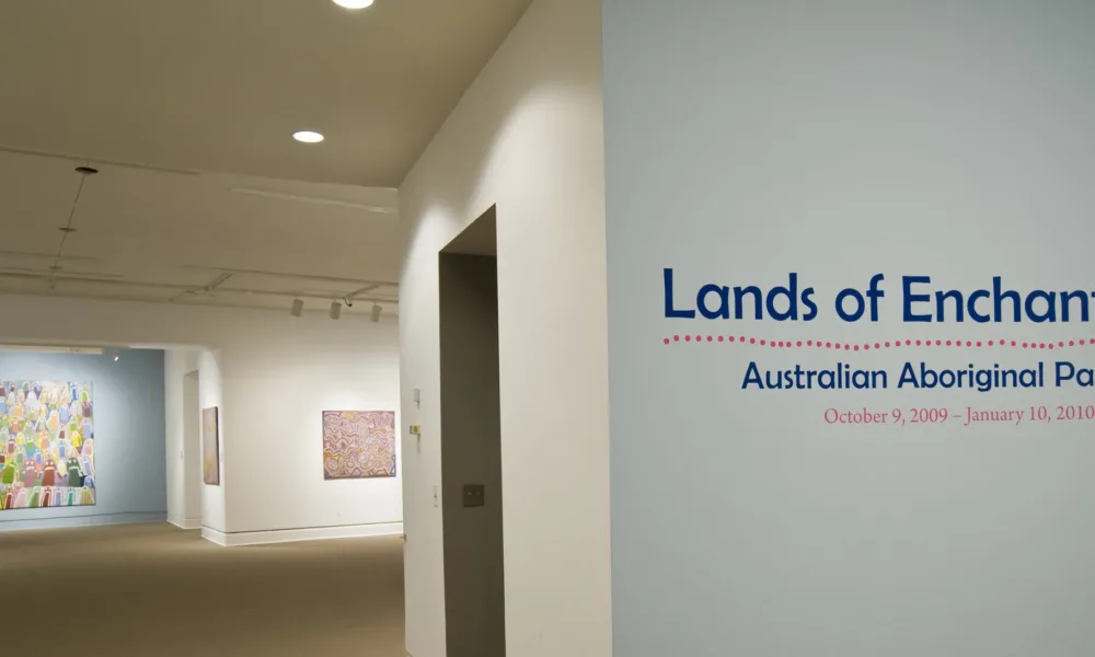 A museum gallery with the words "Lands of Enchantment: Australian Aboriginal Painting Installation" written on one of the walls. There are abstract paintings in the background.