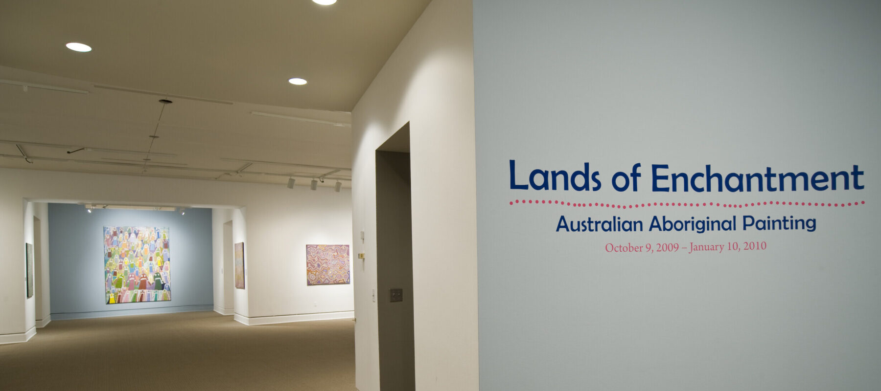 Installation view of a gallery space. On a white wall, it says in big blue letters "Lands of Enchantment: Australian Aboriginal Painting." In the back, there is a large painting hanging on another wall.