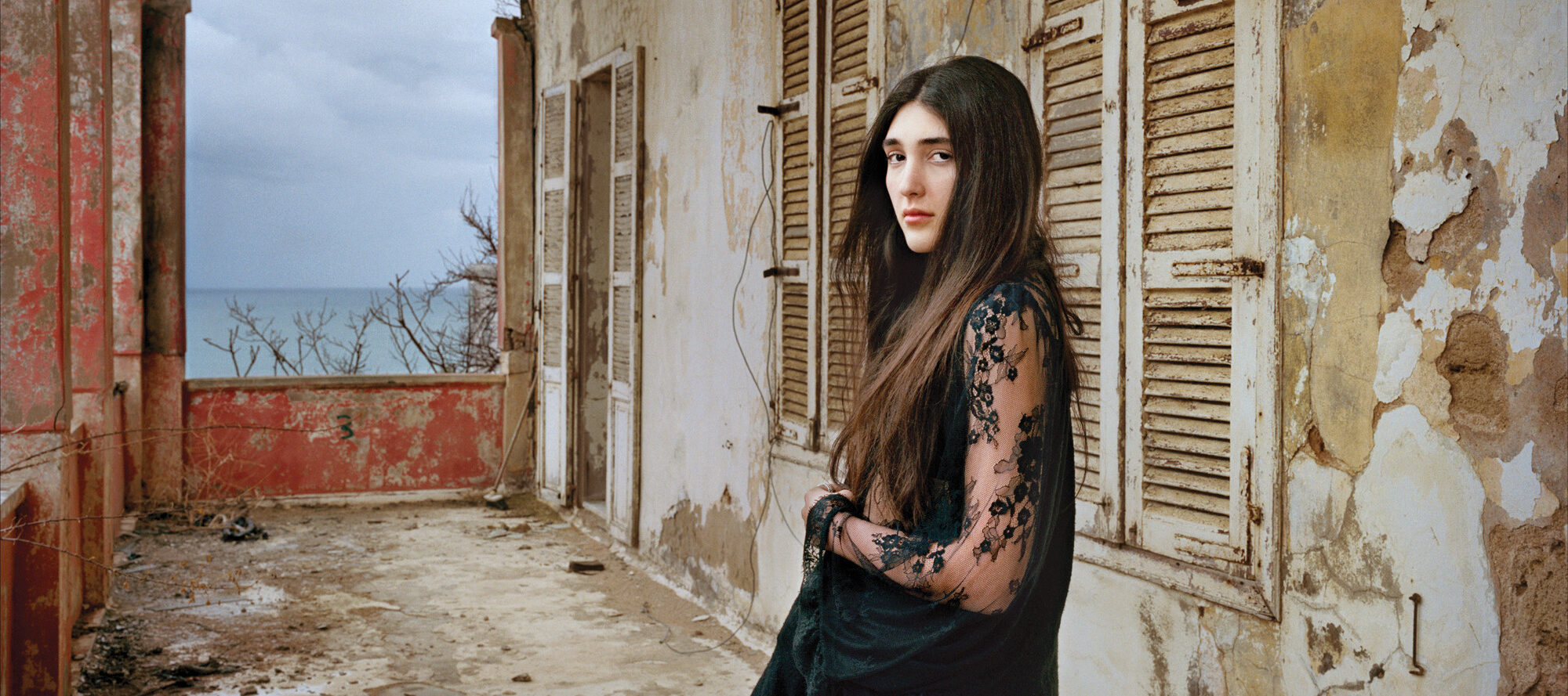 A light-skinned young woman with long, dark brown hair in a black, long lace sleeved dress stands confidently in a crumbling loggia. She gazes at the viewer with a serious, captivating look.