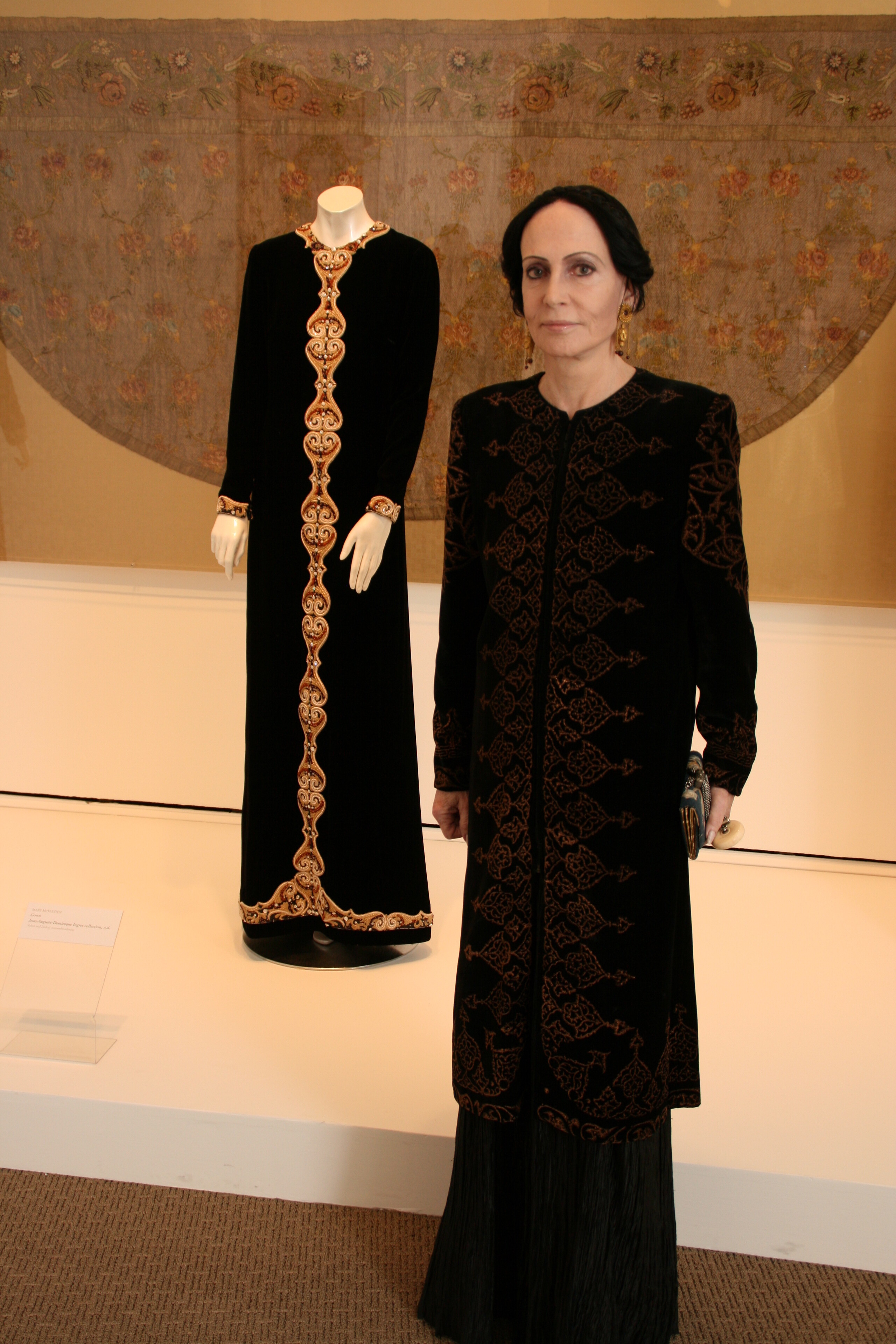 A woman with a light skin tone and black hair in a bun stands before a mannequin wearing a black gown with gold lace.