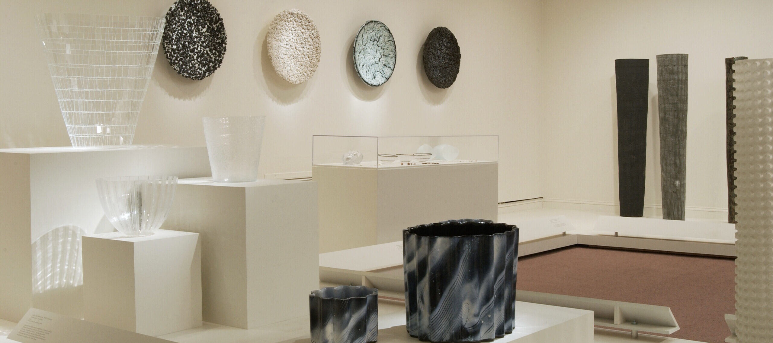 Installation view of a gallery space with white walls and white pedestals. There are several art works on the pedestals, including glass bowls. Several black and gray bowls hang on the wall, as well as some textile art pieces in dark colors.