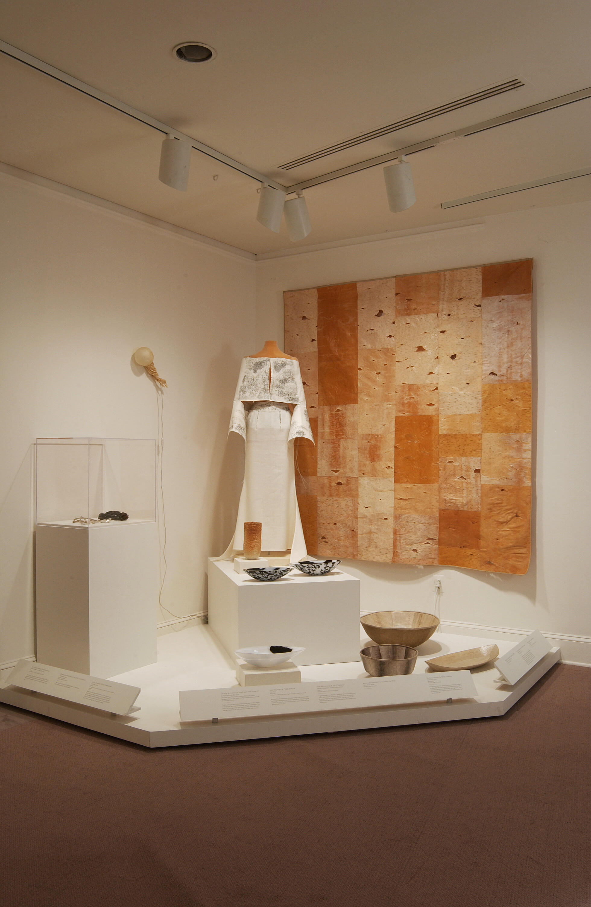 Installation view of a gallery space with white walls and several artworks presented on a pedestal in the corner. The artworks include a large, beige textile art piece hanging from the wall, a mannequin wearing a white costume, and a variety of bowls.