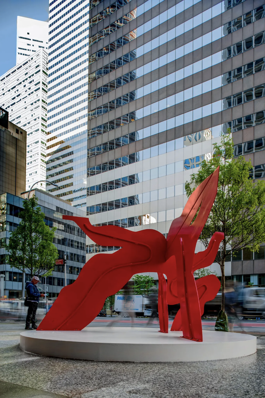 A red sculpture made of metal stands in front of glass highrises. The sculpture’s bright red stands in stark contrast to the plain, gray and black highrise in the background. The sculpture has an organix form, almost like a sea creature it tangles its arms up in the air.