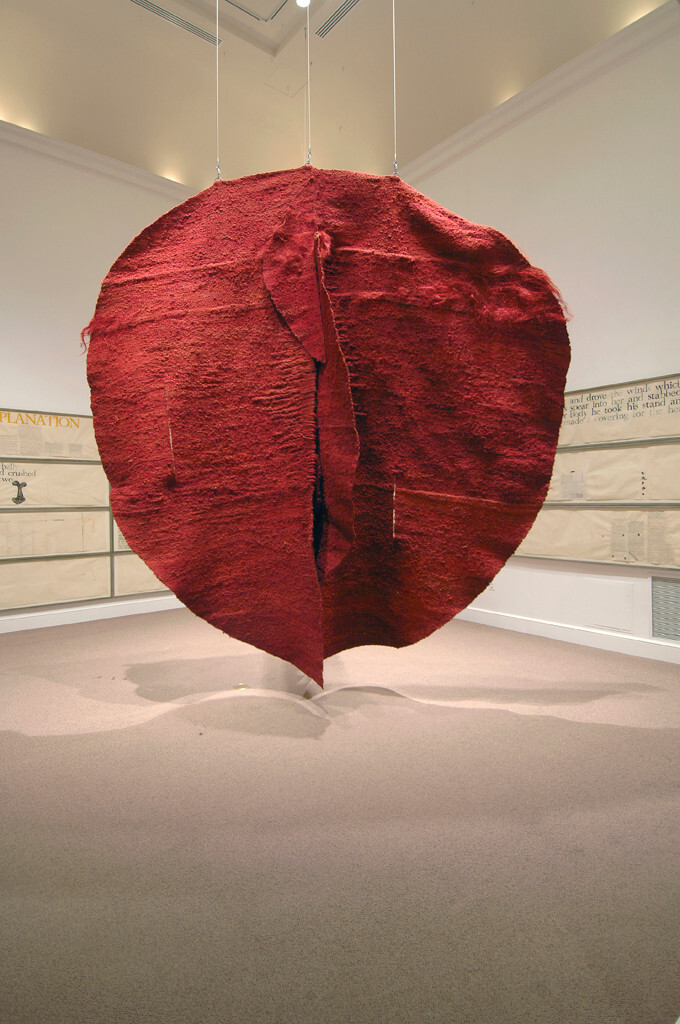 Installation view of a large, textile sculpture hanging from the ceiling in a white gallery space. The textile piece is in a blood-red hue that contrasts with the white walls. The texture is rather rough. It looks like a giant heart hanging from three threads.