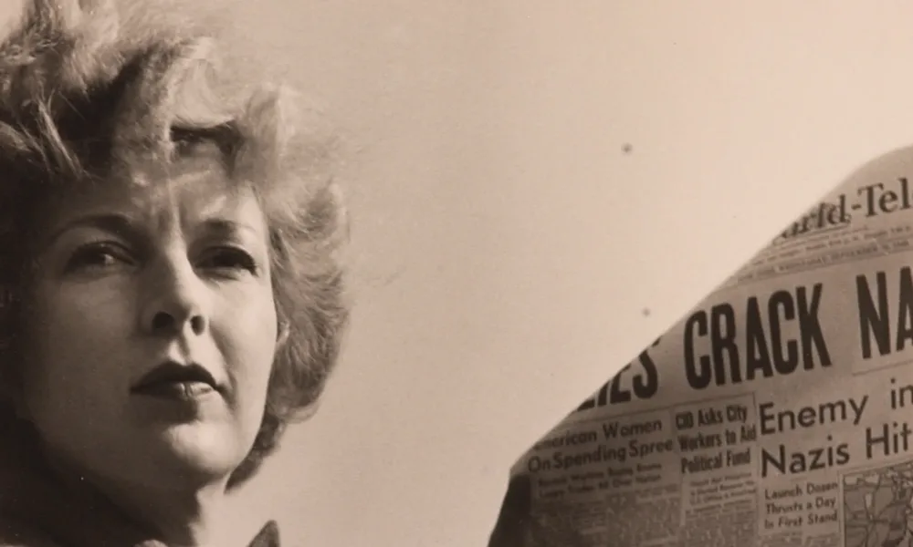 A black-and-white photograph of a light-skinned adult woman holding a newspaper with news about World War II. She wears a coat and her short, curly hair is caught in the wind.