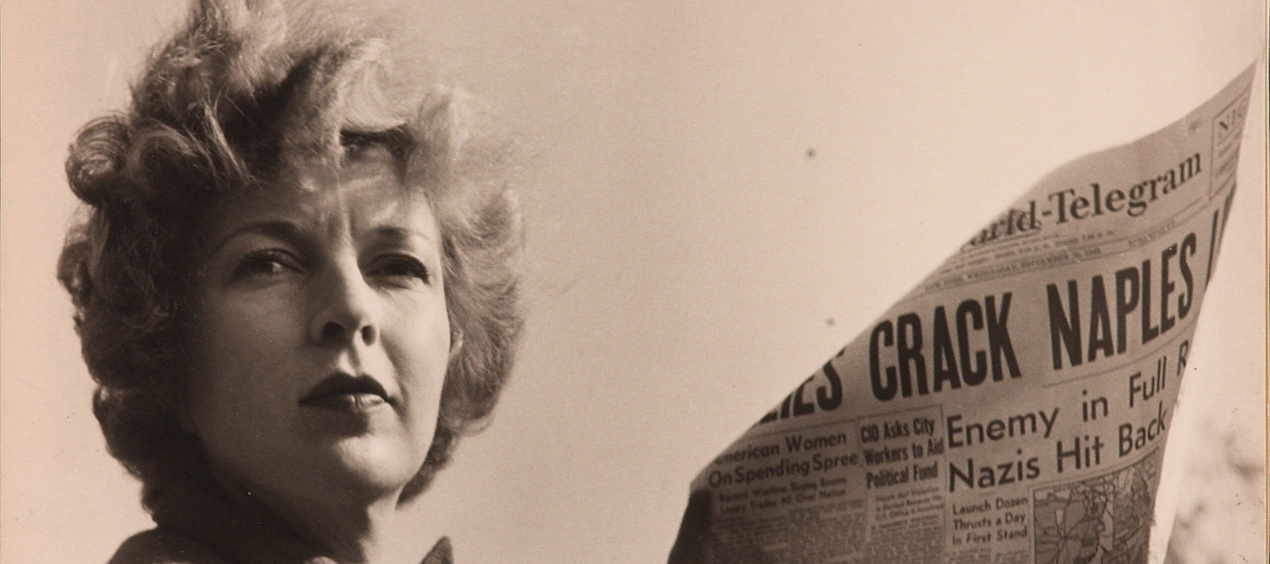 A black-and-white photograph of a light-skinned adult woman holding a newspaper with news about World War II. She wears a coat and her short, curly hair is caught in the wind.