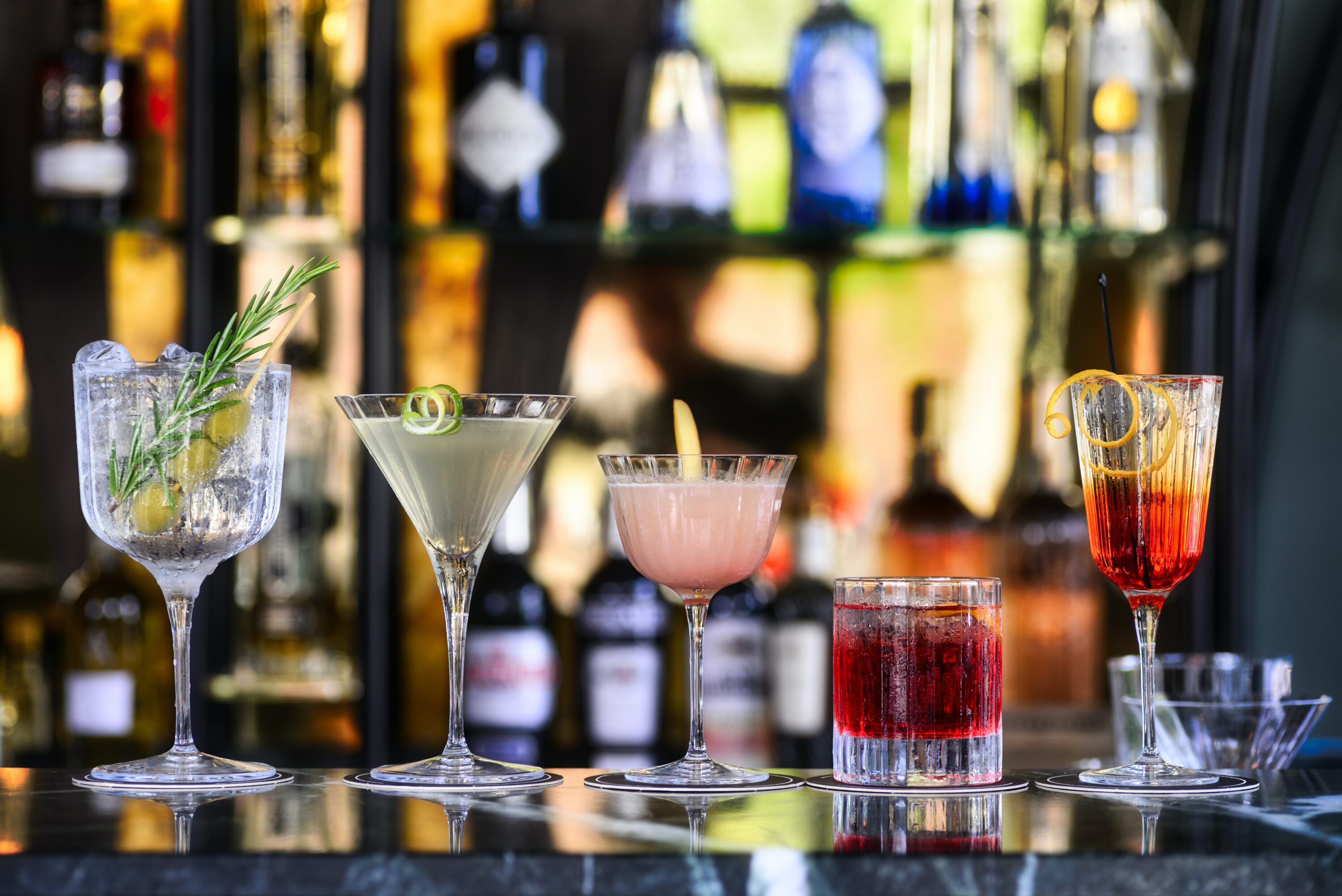 A bar with a line of five colorful cocktails in various shaped glasses.