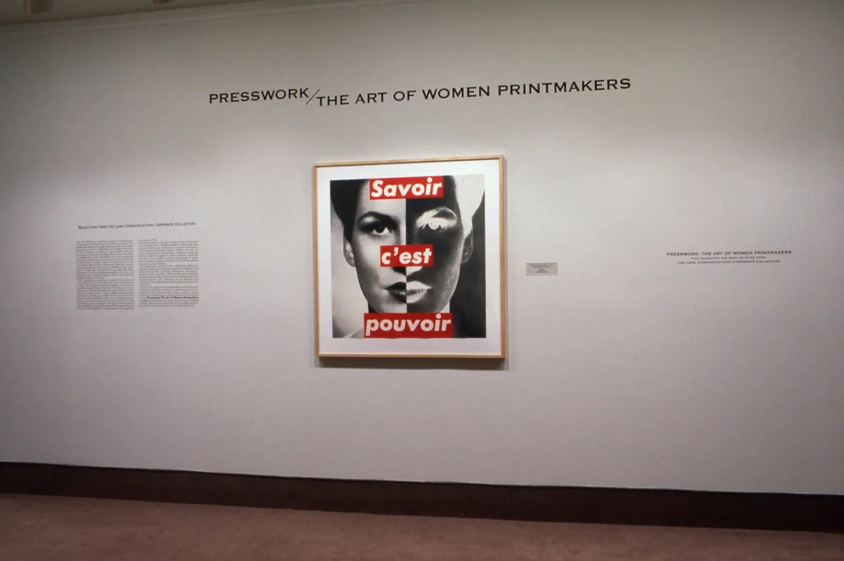 A large photomontage is hanging on a white wall. It's a photograph of a woman's face overlayed with the text "Savoir c'est pouvoir". There is wall text around the artwork, including the title of the exhibition, "Presswork: The Art of Women Printmakers".