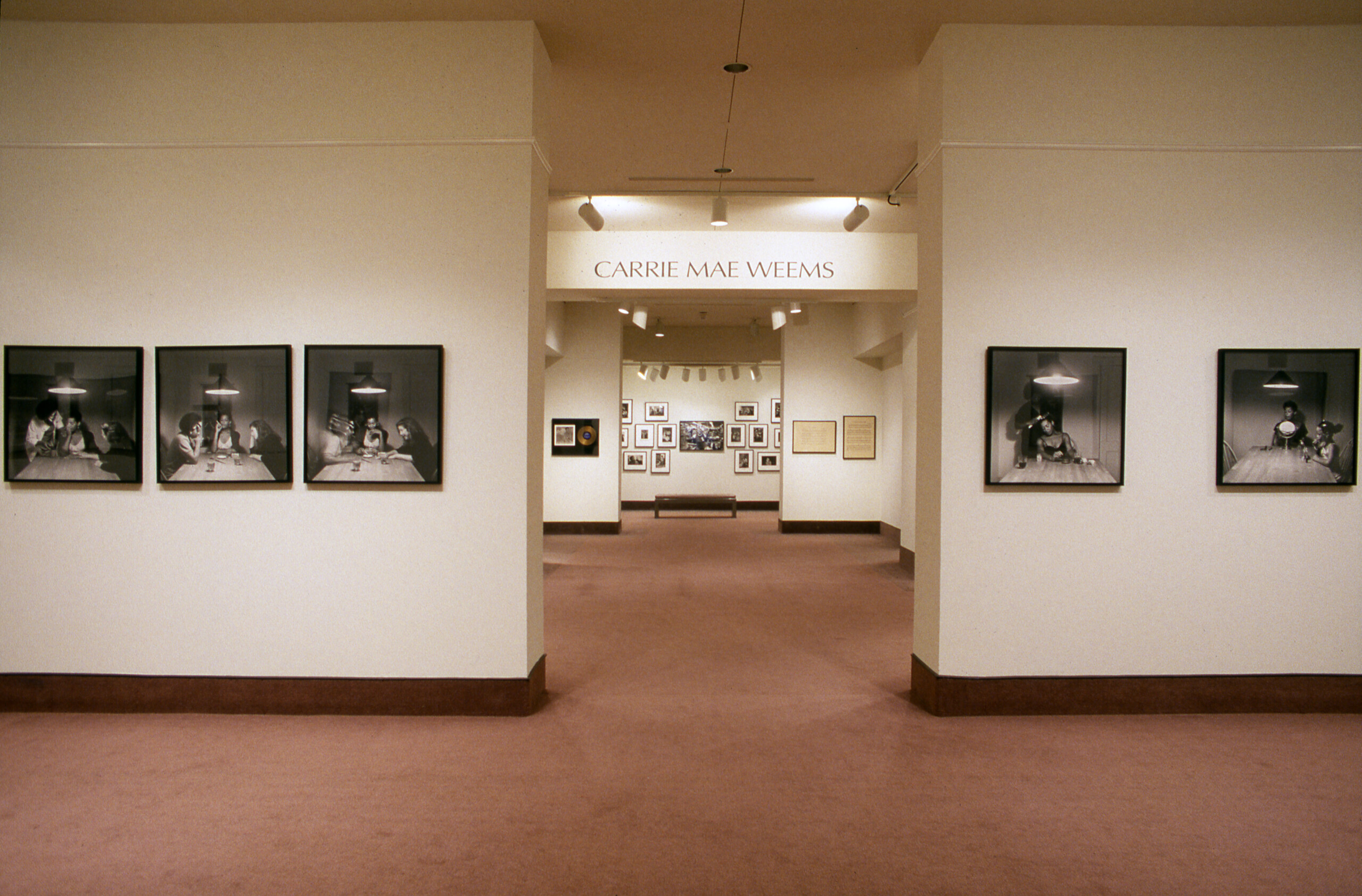 A view of a gallery space. A sequence of photographs of a woman with a dark skin tone sitting around a kitchen table is hanging on the walls. Behind the walls in the back room, it says 