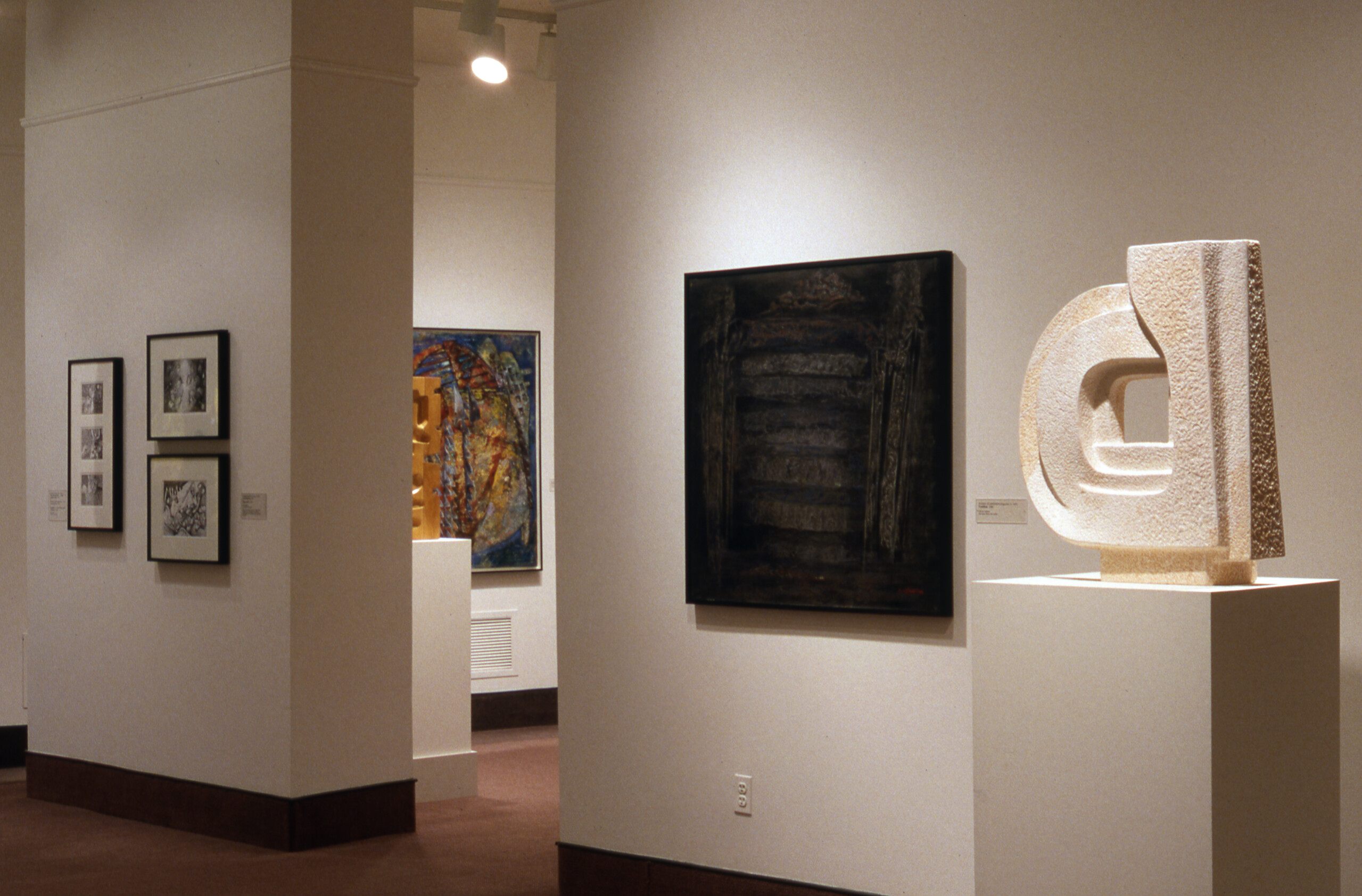A view of a gallery space with several sculptures and paintings. A large white abstract sculpture is standing on the right, right next to an abstract painting in black, juxtaposing each other.