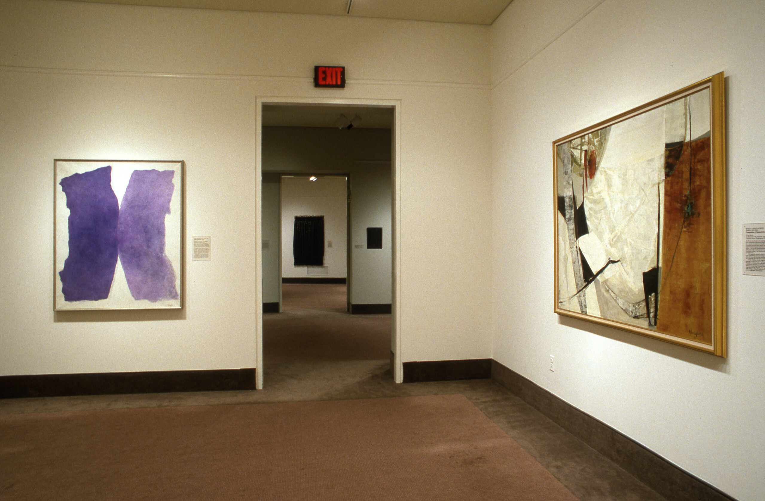 A view of a gallery space. Two artworks are hanging on opposite walls. One is an abstract painting in dark blue, and the other one is an abstract painting in white and brown shades.