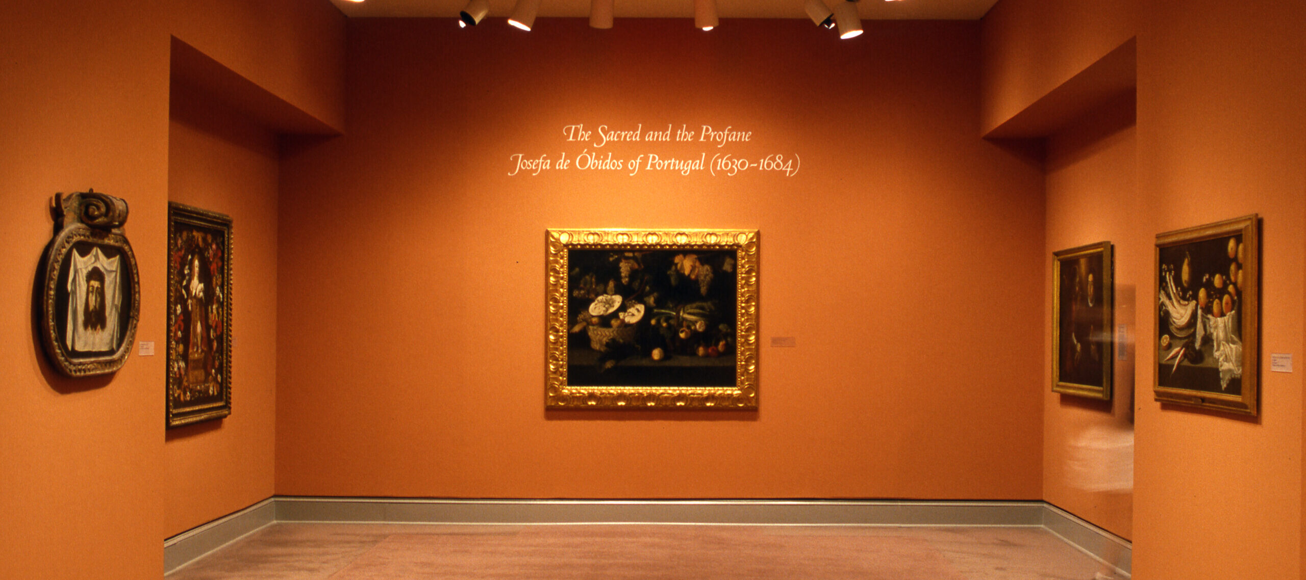 View of a gallery space with orange walls. Several artworks featuring Christian themes in golden frames are hanging on the wall. In the center, a wall text reads: 
