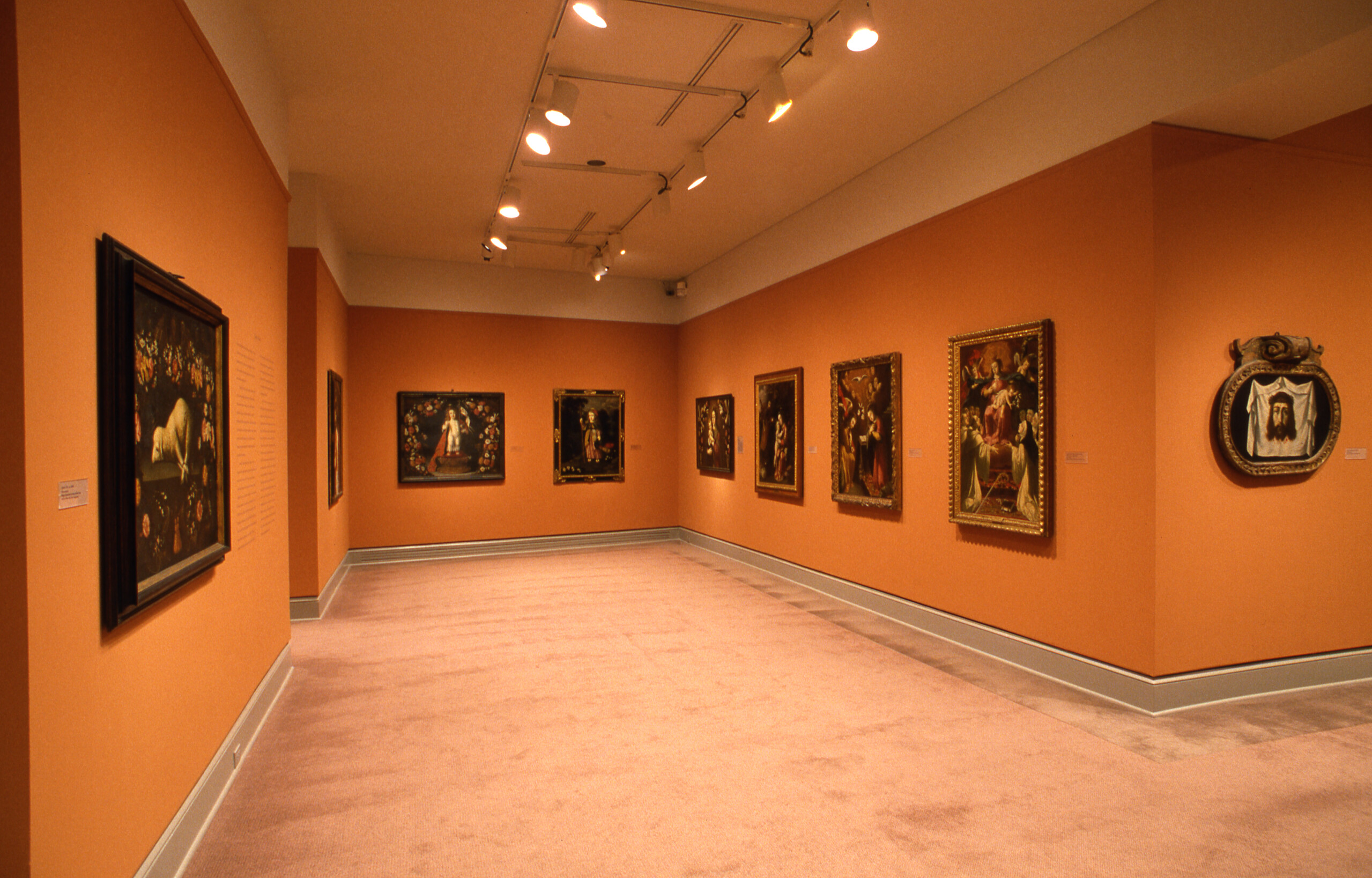 View of a gallery space with orange walls. Several artworks featuring Christian themes in golden frames are hanging on the wall.