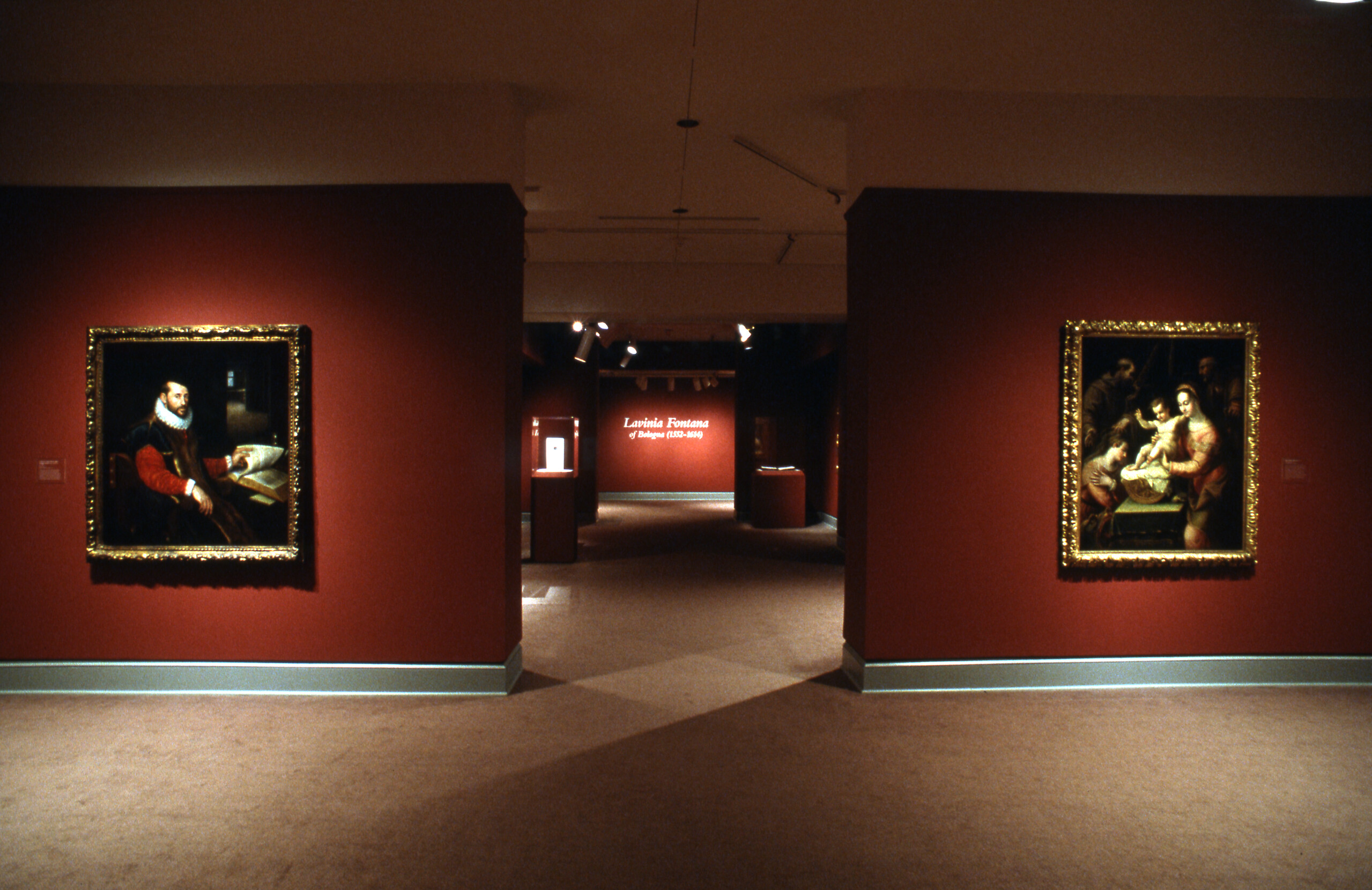 A view of an exhibition space with red walls. Several oil paintings are hanging on opposite walls. They are portraits of men and women in sixteenth-century garments.