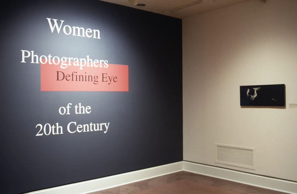 A view of a gallery space. A black-and-white photograph is hanging on one wall. On the opposite wall, it says in large letters: "Defining Eye: Women Photographers of the 20th Century"