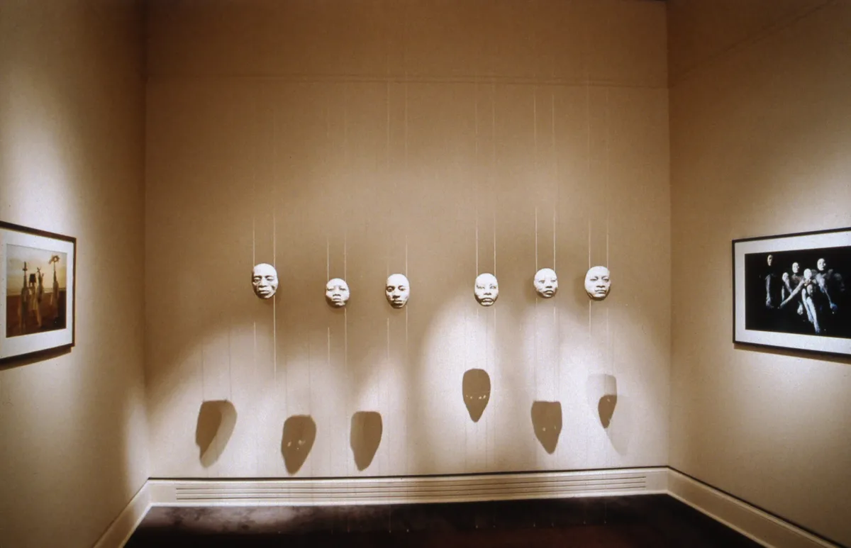 A view of a gallery space with two paintings on opposite walls and six masks hanging down from the ceiling. The masks are white and attached on transparent strings. They cast shadows on the wall behind them, creating a creepy atmosphere.